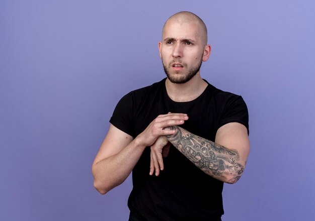 Looking at side thinking young sporty man holding hands together isolated on purple background