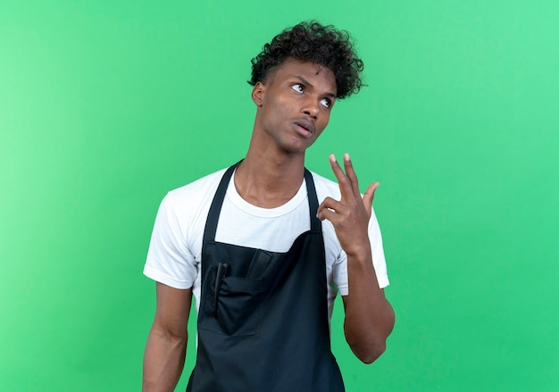 Looking at side tired young afro-american male barber wearing uniform showing pistol gesture isolated on green background