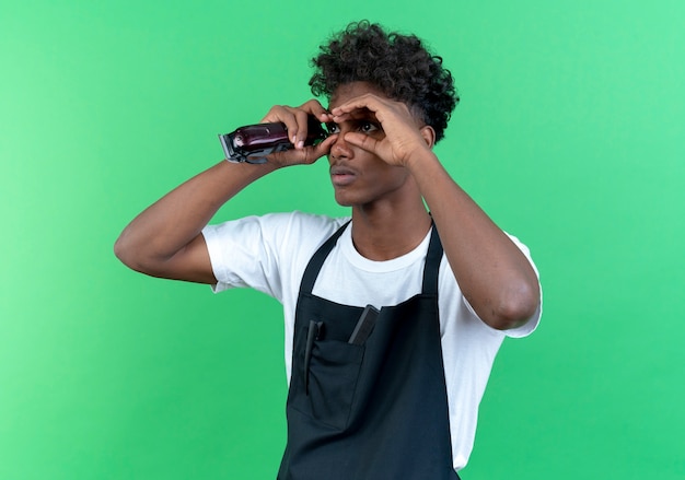 Looking at side young afro-american male barber wearing uniform holding hair clippers and showing look gesture isolated on green background