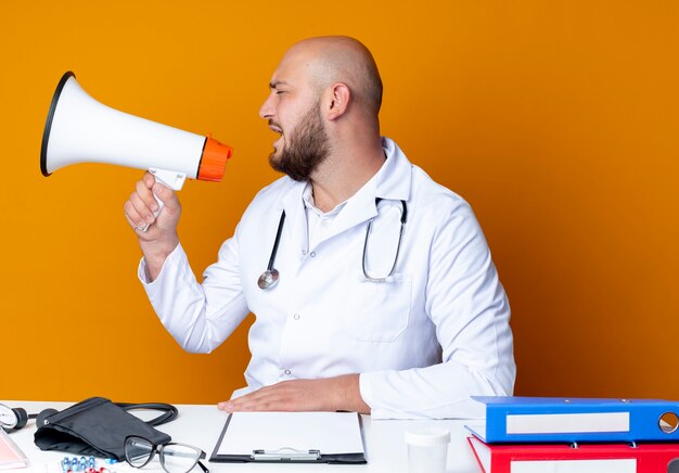 Looking at side young bald male doctor wearing medical robe and stethoscope sitting at work desk with medical tools speaks on loudspeaker isolated on orange background