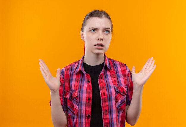 Looking at side young beautiful girl wearing red shirt spreads hands on isolated yellow background