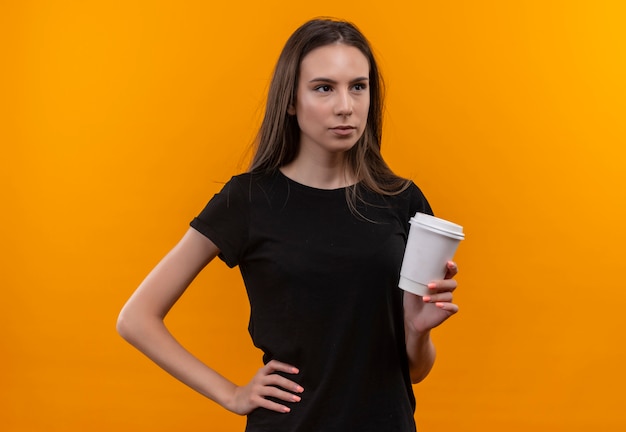 Free photo looking at side young caucasian girl wearing black t-shirt holding cup of coffee put her hand on hip on isolated orange background