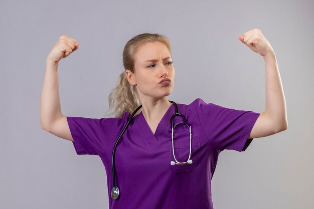 Looking at side young doctor wearing purple medical gown and stethoscope doing strong gesture on isolated white wall