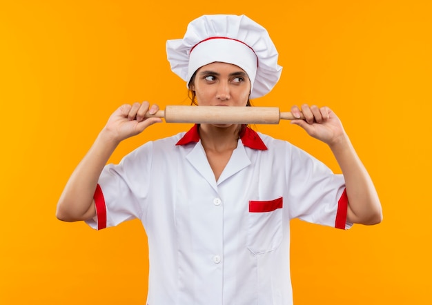 Free Photo looking at side young female cook wearing chef uniform holding and bites rolling pin on isolated yellow wall with copy space