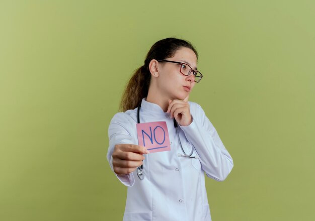 Looking at side young female doctor wearing medical robe and stethoscope with glasses holding out paper note isolated