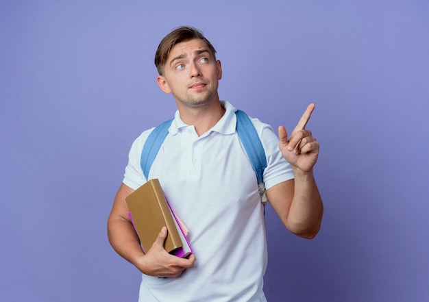 Looking at side young handsome male student wearing back bag holding books and points at side isolated on blue wall with copy space