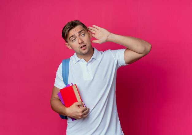 Looking at side young handsome male student wearing back bag holding books and showing listen gesture isolated on pink wall