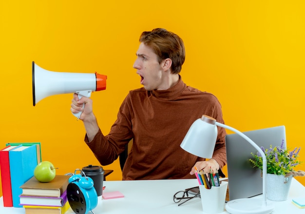 Looking at side young student boy sitting at desk with school tools speaks on loudspeaker 