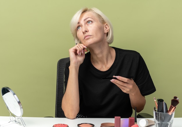 Free photo looking up young beautiful girl sits at table with makeup tools holding makeup brush isolated on olive green wall