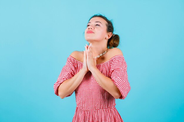 Looking up young girl is wishing by holding hands together under chin on blue background