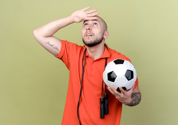 Looking at up young sporty man with jump rope on shoulder holding ball and putting hand on forehead 