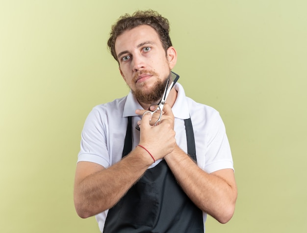 Looking  young male barber wearing uniform holding comb with scissors around face isolated on olive green wall