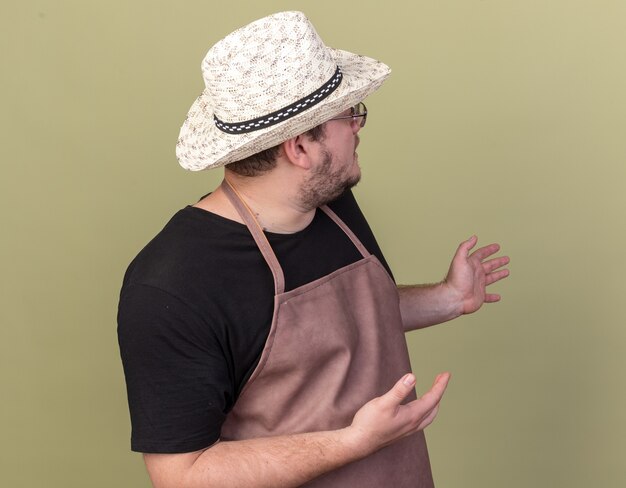 Looking at behind young male gardener wearing gardening hat points at behind isolated on olive green wall with copy space