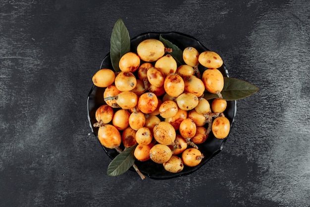 Loquats with leaves in a bowl on dark textured background, top view.