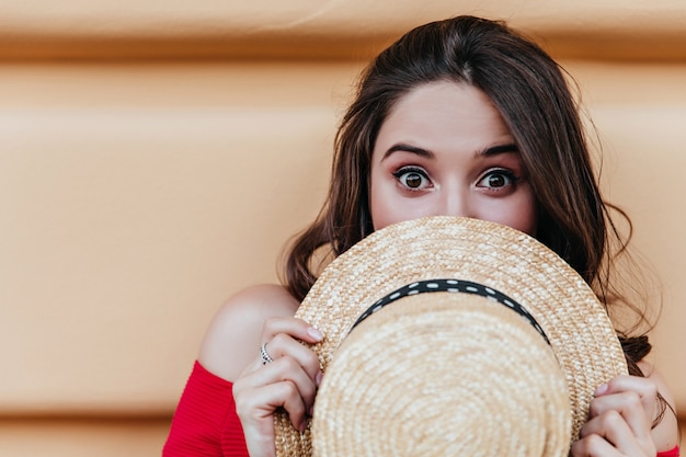 Lovable dark-haired girl posing in front of wall with surprised face expression. Outdoor shot of good-humoured brunette lady hiding behind summer hat.