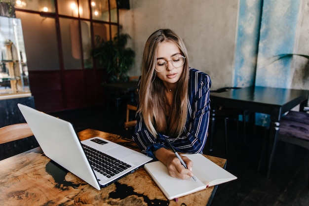 Lovable girl writing something in her notebook. Indoor shot of adorable long-haired woman using white laptop in cafe.