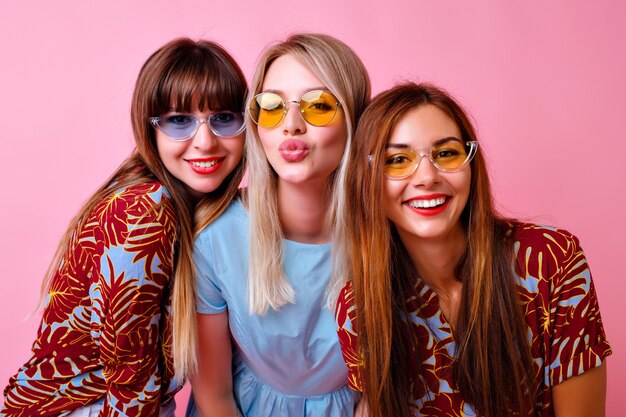 Lovable group of stylish girls smiling and sending kiss, super trendy tropical print clothes and 90s style colorful glasses, best friends enjoy time together, pink wall