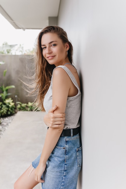 Lovable white female model in denim skirt smiling in morning. Outdoor photo of attractive woman with long brown hair posing near wall.