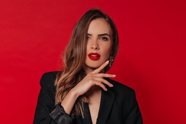 Lovable woman with evening make up posing over red wall, touching her face