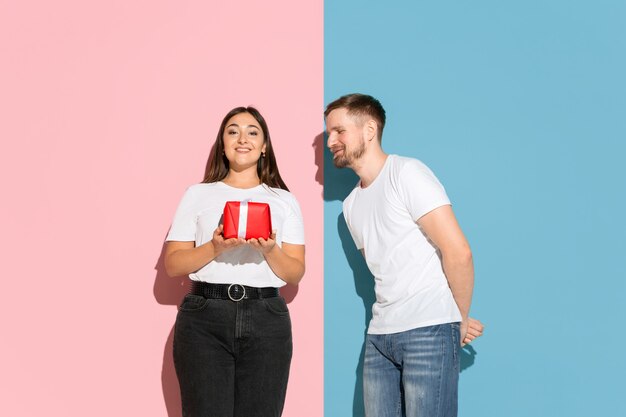 Love. Giving a gift for St. Valentines Day. Young man and woman in casual clothes on pink, blue bicolored wall. Concept of human emotions, facial expession, relations, ad. Beautiful couple.