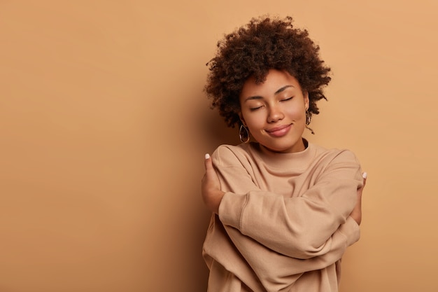 Free photo love yourself, self acceptance. gentle lovely afro american woman crosses hands and embraces own body, tilts head and closes eyes