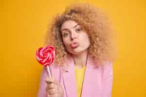 Free photo lovely affectionate woman with curly hair keeps lips folded holds delicious candy on stick has sweet tooth dressed in formal clothes isolated over yellow wall sends air kiss