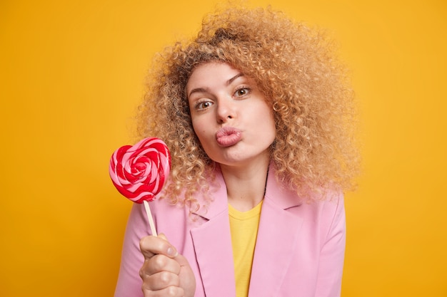 Lovely affectionate woman with curly hair keeps lips folded holds delicious candy on stick has sweet tooth dressed in formal clothes isolated over yellow wall sends air kiss 