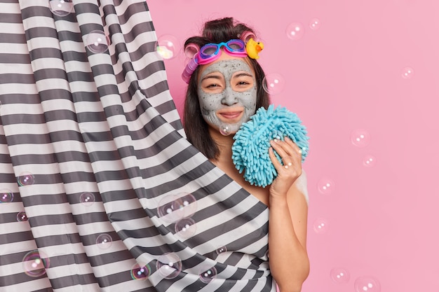 Free Photo lovely brunette woman applies clay mask on face feels refreshed holds bath sponge applies clay mask for skin rejuvenation poses behind shower curtain isolated over pink background with bubbles