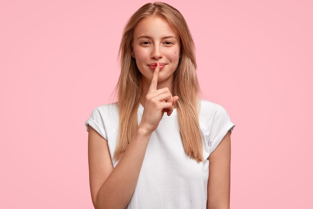 lovely cheerful young European woman touches fore finger on lips, makes silence gesture