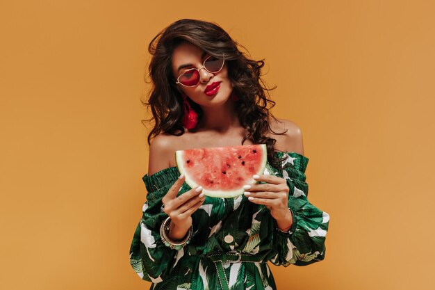 Lovely curly haired girl with red lips and stylish earrings in printed stylish sundress and cool sunglasses holding watermelon