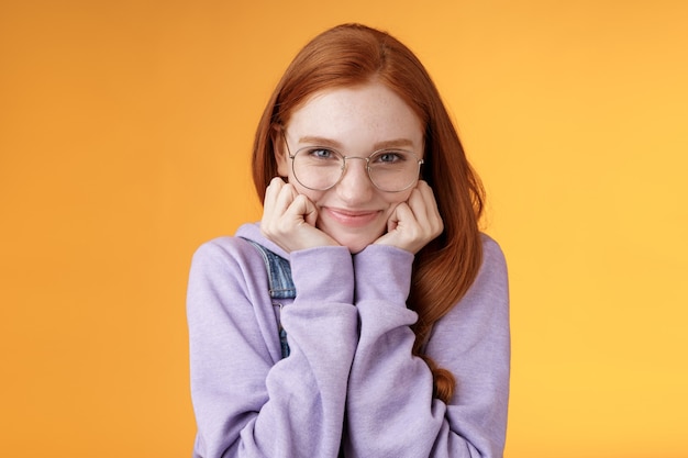 Free photo lovely cute redhead sweet silly girl geek university student wearing glasses lean hand smiling tenderly look affection adore listen sensual confessions boyfriend, standing orange background