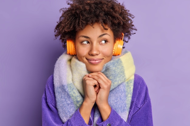 Free Photo lovely dreamy woman with curly hair keeps hands together looks away and thinks about something pleasant listens music via stereo electronic headphones wears warm scarf around neck. hobby concept