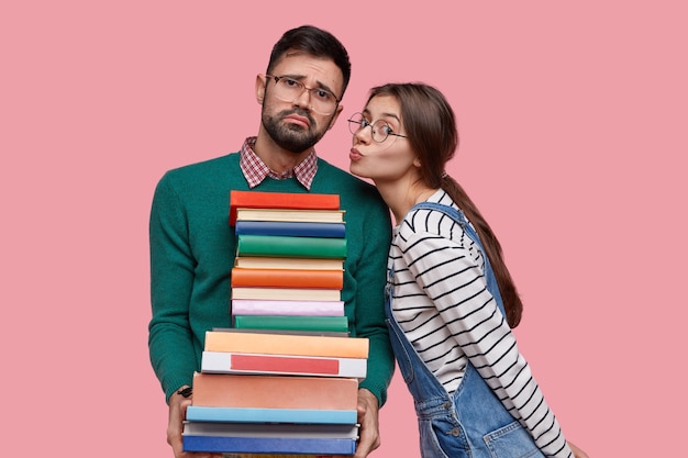 Free photo lovely european schoolgirl going to kiss her boyfriend who holds large pile of books, stand closely to each other