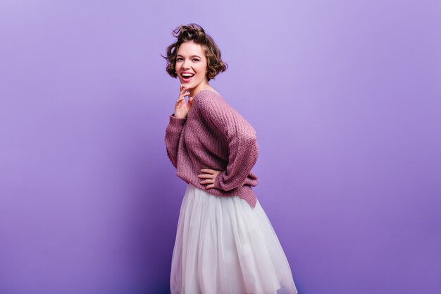 Lovely excited woman with stylish haircut posing in long white skirt.  Indoor photo of joyful brunette female model isolated on bright purple wall.