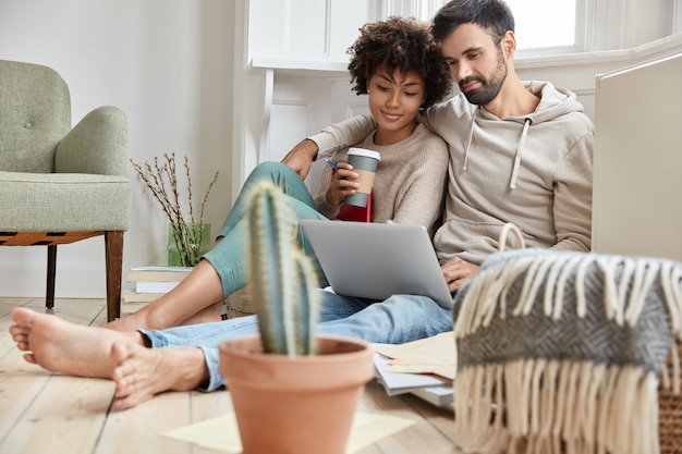 Free photo lovely family couple cuddle together, dressed casually, enjoy domestic atmosphere, synchronize data on laptop computer, work on family business project, drink hot beverage, cactus in foreground