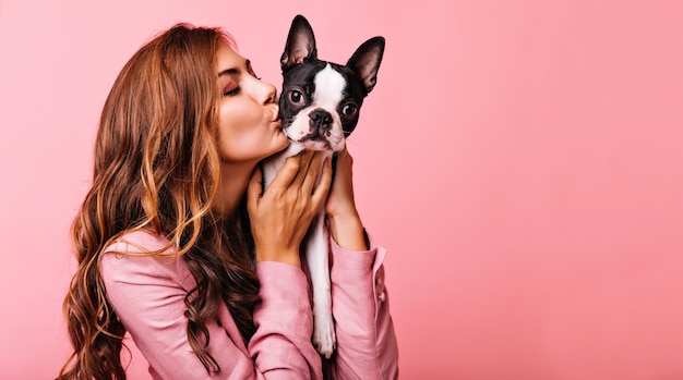 Free photo lovely female model kissing funny bulldog puppy indoor portrait of refined darkhaired girl posing on pink background with dog