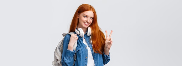 Free photo lovely flirty and cheeky redhead college girl wear headphones over neck and backpack showing peace s