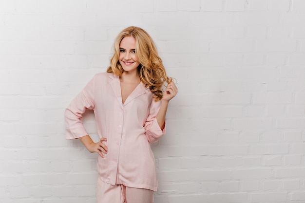 Free photo lovely girl in cotton pyjama playing with her wavy hair.  portrait of laughing woman in pink night-suit smiling on white wall.