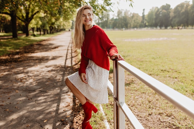 Free photo lovely girl in red nice shoes and white dress enjoying herself in the park. beautiful blonde woman posing playfully near yellow fallen leaves.