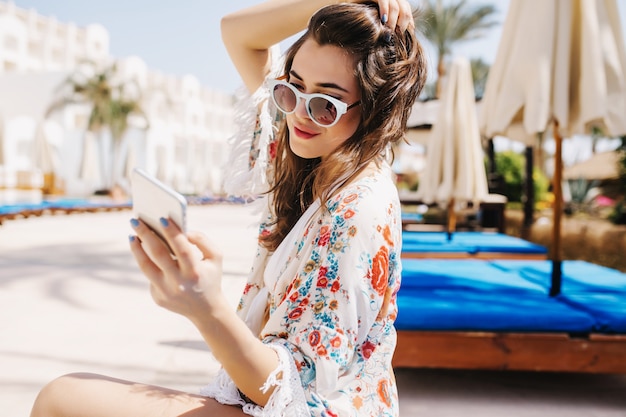 Lovely girl in trendy white sunglasses writing message to friend, holding her dark hair. Portrait of amazing brunette young woman in stylish shirt sitting outside with phone