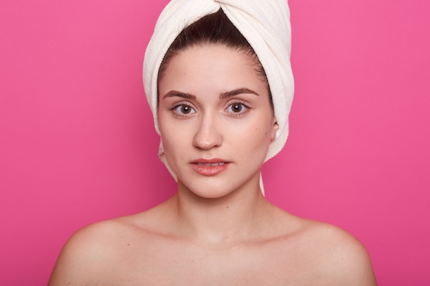 Lovely girl wearing white towel on head, posing with bared shoulders isolated on pink studio, attractive lady looking directly at camera, having clean perfect skin. Skinncare concept.