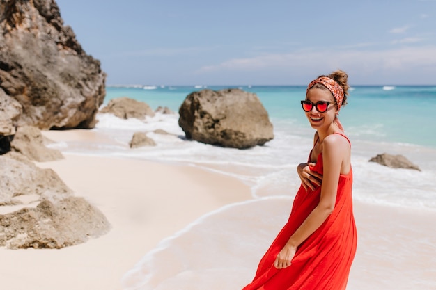 Lovely girl with charming smile spending morning near ocean. Outdoor photo of european young lady in red dress having fun at sea resort.