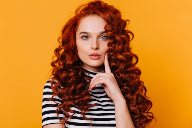 Free photo lovely girl with red curls and blue eyes touches her cheek with her finger and looks thoughtfully at camera on isolated orange space.