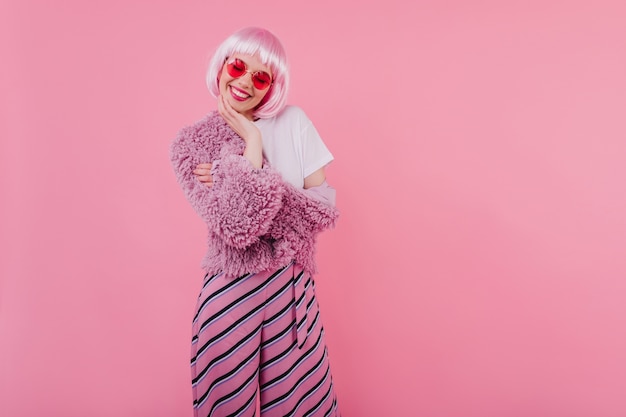 Free photo lovely girl with short pink hair laughing with eyes closed. refined young lady wears peruke and trendy glasses during photoshoot