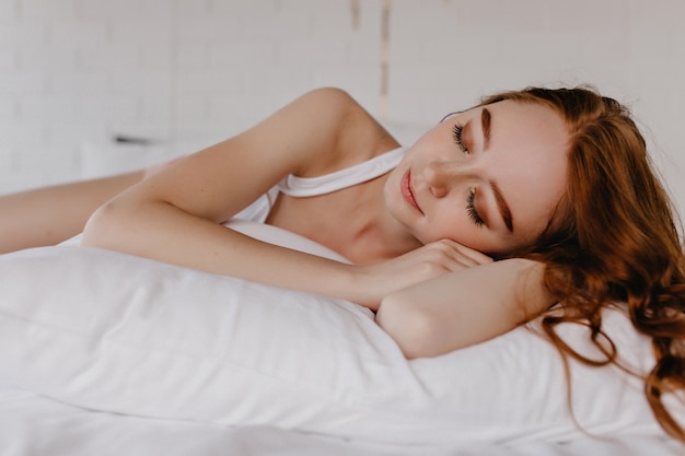 Lovely girl with wavy hairstyle sleeping on pillow. Indoor photo of tired ginger female model with makeup.