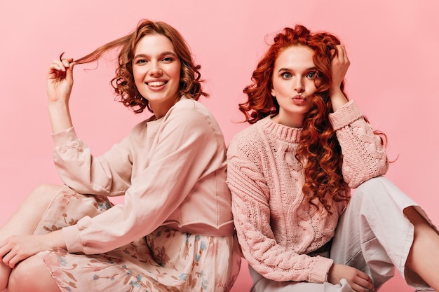 Free photo lovely girls sitting on pink background and touching curly hair. front view of friends posing on floor.