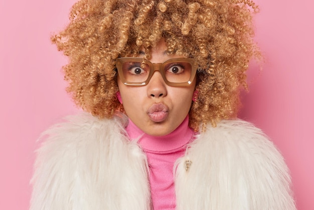 Free Photo lovely good looking woman with curly bushy hair has romantic expression confesses in love keeps lips rounded wears spectacles white fur coat isolated over pink background blows mwah at camera