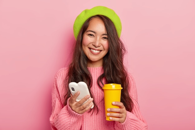 Lovely happy woman with glad expression, uses cellphone for surfing social networks and chatting online, holds yellow takeaway cup with coffee