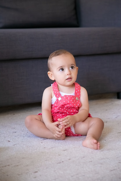 Free photo lovely little baby sitting on carpet barefoot in living room. cute thoughtful girl in red dungarees shorts looking at someone and touching foot. weekend, childhood and being at home concept