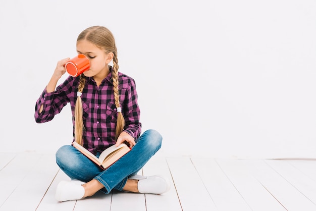 Free photo lovely little girl reading a book and holding a mug